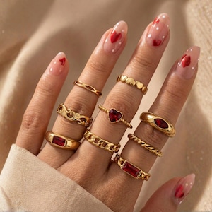 May include: A hand displays a collection of gold rings with red gemstone accents. The rings feature various designs, including a heart-shaped stone, a chain link style, and a band with a rectangular stone. The nails are painted with a clear base and red heart designs.