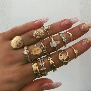 May include: A collection of gold-toned rings of various designs, including a coin ring, floral, and gemstone accents. The rings are displayed on a hand with glittery nail polish, showcasing a bohemian jewelry style.