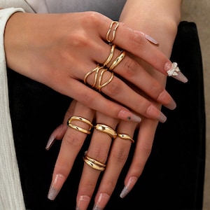 Puede incluir: Una colección de anillos dorados de varios diseños, incluyendo una doble banda, un diseño ondulado y una banda simple. Los anillos se muestran en una mano con uñas largas y cuidadas. El tema general es la moda y la joyería.