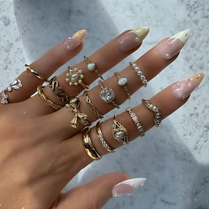 Puede incluir: Una mano que muestra una colección de anillos de oro, incluyendo bandas, diseños ornamentados y anillos con piedras preciosas y perlas. Las uñas son largas, con manicura francesa y detalles decorativos.