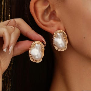 Peut inclure: Une paire de boucles d'oreilles dorées présentant un centre en fausse perle blanche de forme organique irrégulière, serti dans un cadre en métal texturé au style fondu. Les boucles d'oreilles ont un fermoir à tige et une finition polie.