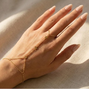 Puede incluir: Una cadena de mano dorada con abalorios en forma de estrella. Esta delicada joya se conecta a un anillo y se envuelve alrededor de la muñeca. La cadena de mano se muestra sobre un fondo de tela clara.