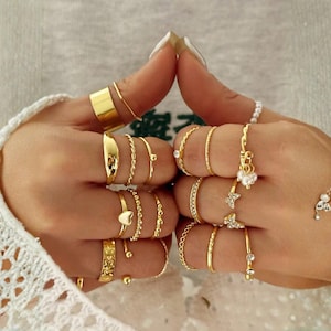 Puede incluir: Dos manos adornadas con una variedad de anillos dorados. Los anillos presentan diversos diseños, incluyendo bandas lisas, patrones trenzados y anillos con pequeños elementos decorativos. También se observa una pulsera de perlas y otra con detalles florales.
