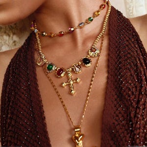 Puede incluir: Un conjunto de collares dorados en capas. El collar superior tiene pequeñas piedras preciosas de colores. El collar del medio tiene piedras preciosas de colores más grandes y un colgante de cruz. El collar inferior tiene un colgante en forma de corazón.