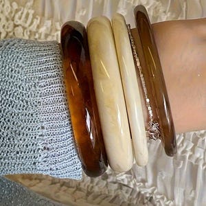 Może przedstawiać: Biały ceramiczny kubek z czarnym uchwytem i napisem "But First, Coffee" pogrubioną czcionką. Kubek jest umieszczony na rozmytym tle, sugerującym przytulne otoczenie.