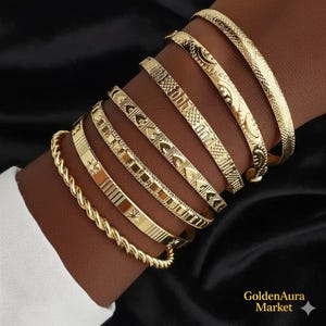 Puede incluir: Una colección de brazaletes dorados con varios diseños, incluyendo estilos texturizados, estampados y trenzados. Los brazaletes se muestran en un brazo sobre un fondo negro. El logotipo de GoldenAura Market es visible en la esquina inferior derecha.