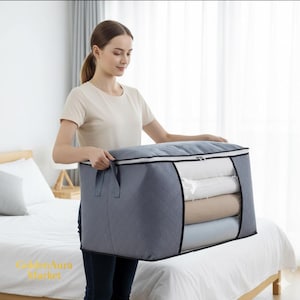 Puede incluir: Una mujer sostiene una bolsa de almacenamiento gris grande con un patrón acolchado. La bolsa tiene una ventana transparente que muestra ropa de cama doblada en el interior. La bolsa tiene cierre de cremallera y asas para transportar. La bolsa está sobre una cama.