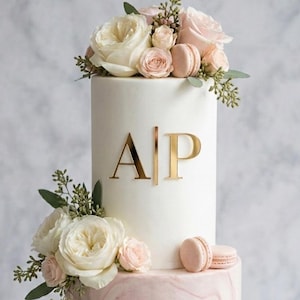 Könnte beinhalten: Eine mehrstöckige Torte mit weißem Zuckerguss, verziert mit goldenen Initialen "A" und "P". Die Torte ist mit frischen Rosen, Grünzeug und Macarons dekoriert. Die untere Etage hat ein rosa Marmormuster.