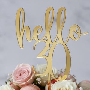 Könnte beinhalten: Ein goldfarbener Tortenaufsatz mit dem Schriftzug "hello 30" in Schreibschrift. Der Aufsatz befindet sich auf einem Blumengesteck mit rosa und weißen Rosen. Der Hintergrund ist ein weiches, verschwommenes Grau.