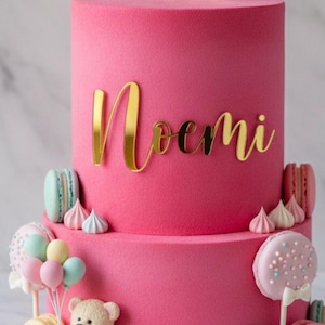 Könnte beinhalten: Ein zweistöckiger rosa Kuchen mit goldenen Buchstaben, die "Noemi" buchstabieren. Der Kuchen ist mit Macarons, Baisers, einem Teddybär und ballonförmigen Lollis verziert. Der Kuchen hat eine glatte, samtige Textur.