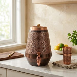 May include: A copper-colored beverage dispenser with an ornate floral pattern. The dispenser has a spigot and a lid with a gold-colored knob. Two matching copper cups are stacked on a linen napkin. A wooden cutting board and a bowl of fruit are also visible.