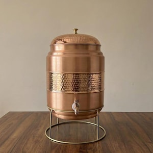 Puede incluir: Un dispensador de agua de cobre con una banda de textura martillada y un grifo. El dispensador tiene una tapa con un pequeño pomo y se apoya en un soporte de metal dorado. El dispensador está sobre una mesa de madera.