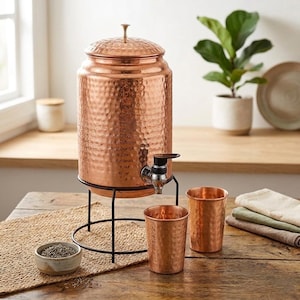 May include: A copper water dispenser with a hammered texture, a spigot, and a lid, set on a black metal stand. Two matching copper cups and a small bowl are on a wooden table. A plant in a white pot is in the background.