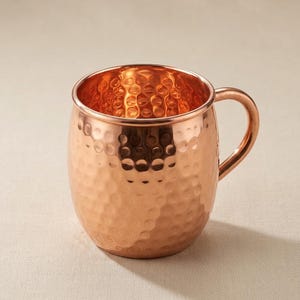 May include: A copper Moscow Mule mug with a hammered finish. The mug has a rounded body, a handle, and a shiny interior. The copper colour is a warm, reddish-brown hue. The mug is designed for serving cold beverages.