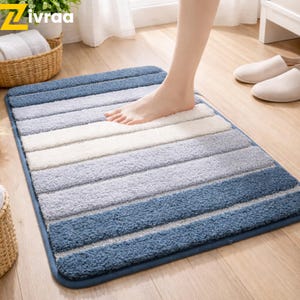 Puede incluir: Una alfombra de baño rectangular con rayas horizontales en tonos azules, grises y blancos. La alfombra tiene un borde azul oscuro. Un pie está colocado sobre la alfombra. La alfombra está sobre un suelo de madera.