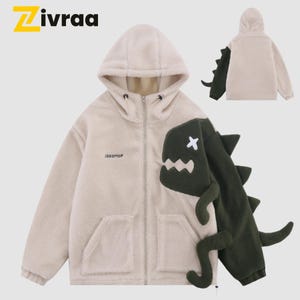 Puede incluir: Chaqueta de forro polar beige con un diseño de dinosaurio en la parte delantera y la manga. La chaqueta tiene capucha, cierre de cremallera y dos bolsillos delanteros. El dinosaurio es verde oscuro con una cruz blanca para un ojo. La marca "Zivraa" está arriba a la izquierda.