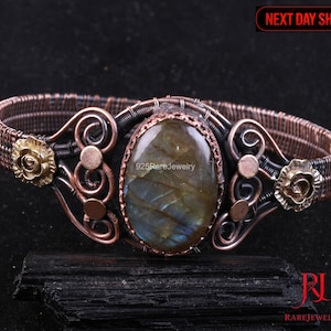 May include: A handcrafted copper wire bracelet with a large oval labradorite stone at the center. The bracelet features decorative swirls, brass accents, and small brass rose details. The text "925RareJewelry" is visible.