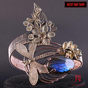 Op de afbeelding: Handgemaakte koperen en messing manchetarmband met een libelle, bloemaccenten en een blauwe labradorietsteen. De armband heeft een uniek, artistiek ontwerp met ingewikkeld draadwerk en een op de natuur geïnspireerd thema.