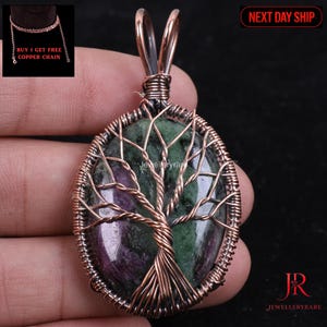 Puede incluir: Colgante ovalado con una piedra verde y morada, intrincadamente envuelto con alambre de cobre en un diseño de árbol de la vida. El colgante incluye una fianza de cobre y se muestra sobre un fondo oscuro. El texto "Jewelleryrare" es visible.