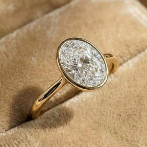 Könnte beinhalten: Ein goldener Ring mit einem großen, oval geformten Diamanten. Der Diamant ist in einer Zargenfassung gefasst, und das Band ist aus poliertem Gold. Der Ring wird auf einer beigen Samtoberfläche präsentiert.
