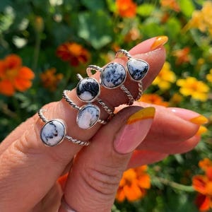 Può includere: Vengono mostrati diversi anelli d'argento con pietre a motivi bianchi e neri. Gli anelli hanno un design a fascia intrecciata. Lo sfondo presenta fiori arancioni e gialli sfocati.