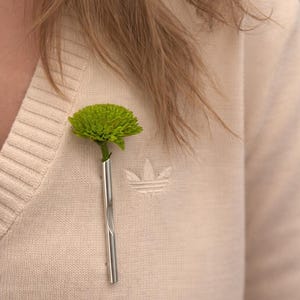 May include: A bright green flower in a silver vase pinned to a cream-coloured sweater. The sweater has a small logo on the right side. The flower contrasts with the neutral tones of the clothing.