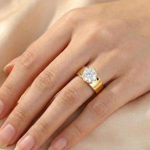 May include: A gold-colored ring with a large, round, clear gemstone. The ring has a wide band and is set on a hand with a neutral background. The gemstone is the focal point, reflecting light and adding sparkle to the jewelry.