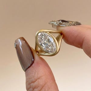 May include: A gold ring featuring a large, oval-cut diamond. The diamond is set in a bezel setting, and the band has a unique, open design. The ring is held up against a neutral background, showcasing its sparkle and craftsmanship.