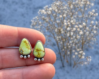 May include: A pair of handcrafted stud earrings. Each earring features a light green, teardrop-shaped stone with brown veining, set in a silver-colored metal. The earrings have a decorative silver-colored base with three small, round accents.