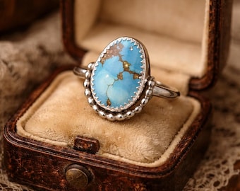 May include: A silver ring with an oval turquoise stone, set in a decorative bezel. The ring is displayed in a vintage brown leather jewelry box with a velvet interior. The turquoise stone has a light blue color with brown veining.