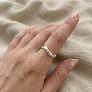Puede incluir: Un anillo plateado con un diseño ondulado. El anillo se lleva en un dedo, mostrando su forma suave y curvada. El diseño del anillo es simple y elegante, reflejando la luz.