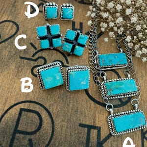May include: A collection of turquoise jewelry pieces, including a necklace and various earring styles. The necklace features three rectangular turquoise stones. Earrings include square and cross-shaped designs, all with silver-toned metalwork. The jewelry is displayed on a wooden surface.