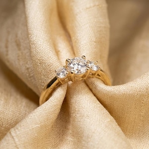 May include: A gold ring with three clear gemstones, set against a backdrop of beige fabric. The ring's band is a polished gold, and the gemstones sparkle. The fabric is draped, creating soft folds and shadows.