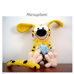 Peut inclure: Une poupée Marsupilami au crochet, jaune avec des taches noires, de grandes oreilles et une longue queue. Le jouet tient un petit jouet bleu. Le texte "Marsupilami" est au-dessus de la poupée.