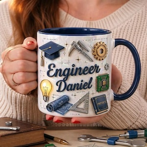 May include: White ceramic mug with a blue handle and rim, decorated with engineering-themed graphics and the name "Engineer Daniel." Features include a graduation cap, gears, and a lightbulb. The mug is held by a person.