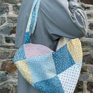 Peut inclure: Un sac fourre-tout en patchwork en forme de cœur avec des motifs floraux bleus, roses et jaunes. Le sac a une bandoulière assortie et est porté à l'épaule. Le sac est en tissu matelassé.