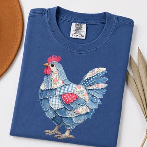 Może przedstawiać: Niebieska koszulka Comfort Colors z patchworkowym wzorem koguta. Kogut wykonany jest z różnych niebieskich tkanin we wzory, w tym dżinsu, kraty i nadruków kwiatowych. Koszulka ma okrągły dekolt i małą metkę z napisem "Comfort Colors".