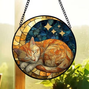 May include: Round stained glass window art featuring a sleeping orange tabby cat. The cat is curled up in a crescent moon shape, with a starry night sky background. The window is framed in black and hangs from a chain.