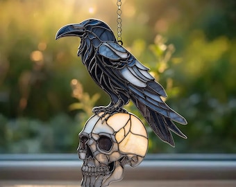 May include: A stained glass raven perched atop a skull, suspended by a chain. The raven is primarily black and gray, with a detailed wing design. The skull is white and cream, with visible bone structure. The background is blurred, with natural light.