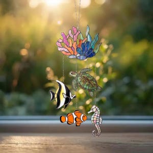 May include: A colorful stained glass mobile featuring coral, a sea turtle, a black and white fish, an orange clownfish, and a seahorse. The mobile is suspended by chains and hangs in front of a blurred green background.
