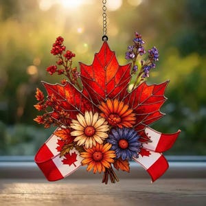 Peut inclure: Ornement décoratif représentant une feuille d'érable rouge avec un bouquet de fleurs colorées et un ruban aux couleurs du drapeau canadien. L'ornement est suspendu par une chaîne.