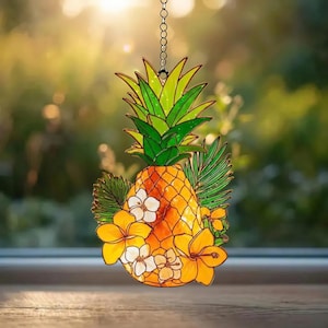May include: A stained glass suncatcher shaped like a pineapple with green leaves, a textured orange body, and yellow and white hibiscus flowers. The piece is suspended by a silver metal chain and features dark lead lines outlining each segment.