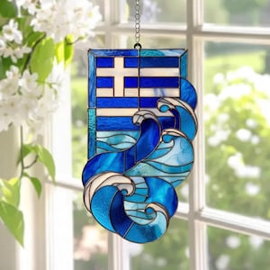 Könnte beinhalten: Glasfenster-Hänger mit dem Design der griechischen Flagge, mit einem weißen Kreuz auf einem blau-weiß gestreiften Hintergrund. Unter der Flagge befinden sich stilisierte blaue und weiße Wellen. Der Fensterhänger wird von einer Kette gehalten.