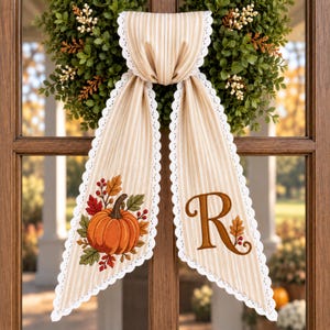 May include: A beige and white striped fabric door hanger featuring a scalloped white lace trim. One side displays an embroidered orange pumpkin with autumn leaves and berries, while the other side features a large brown letter R with a small leaf.