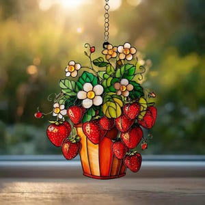 May include: A stained glass suncatcher featuring a cluster of bright red strawberries and white flowers with yellow centres, set in a striped orange and yellow pot. The piece is suspended by a metal chain, showcasing vibrant green leaves.