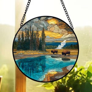 Op de afbeelding: Rond gebrandschilderd glaspaneel met een landschap met bizons, een meer en bomen. Het kunstwerk heeft een levendig kleurenpalet van blauw, groen, oranje en bruin, met een zwarte metalen ketting om op te hangen. De scène omvat een zonsondergang en wolken.