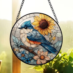 Puede incluir: Una vidriera redonda que representa un pájaro azul posado en una rama con flores y un girasol. El pájaro tiene plumas azules, blancas y naranjas. La ventana está enmarcada en negro y cuelga de una cadena negra.
