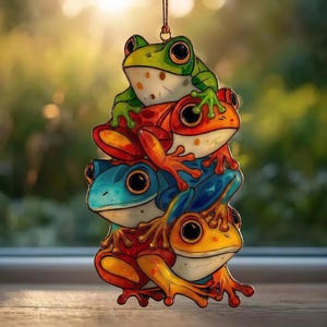 May include: A colorful ornament featuring a stack of four cartoon frogs. The frogs are in shades of green, orange, blue, and yellow, with large black eyes. The ornament hangs from a brown string, perfect for decorating.