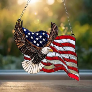 Könnte beinhalten: Ein Buntglas-Suncatcher mit einem Weißkopfseeadler mit braunen und weißen Federn im Flug, vor einem rot-weiß-blauen amerikanischen Flaggendesign. Das Stück ist an einer dünnen Metallkette befestigt, um es an einem Fenster aufzuhängen.