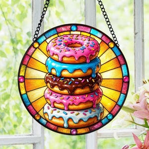 Op de afbeelding: Een rond gebrandschilderd glazen kunstwerk met een stapel van vijf kleurrijke donuts. Elke donut heeft een andere kleur glazuur: roze, blauw, bruin, roze en lichtblauw. De donuts zijn versierd met hagelslag. Het raam heeft een zwarte ketting om op te hangen.